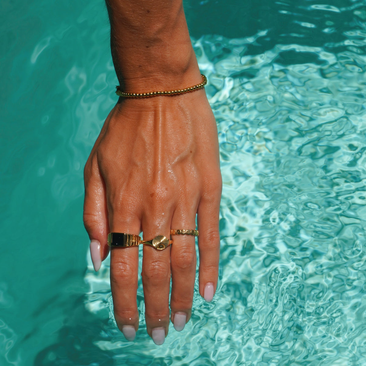 Hand in pool water wearing gold rings and a gold beaded bracelet